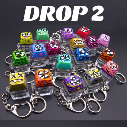 A group of small colorful cube-shaped keychains with cartoon monster faces, each mounted on a clear plastic stand and attached to a silver metal key ring. Large white text at the top reads 'DROP 2' on a dark background.
