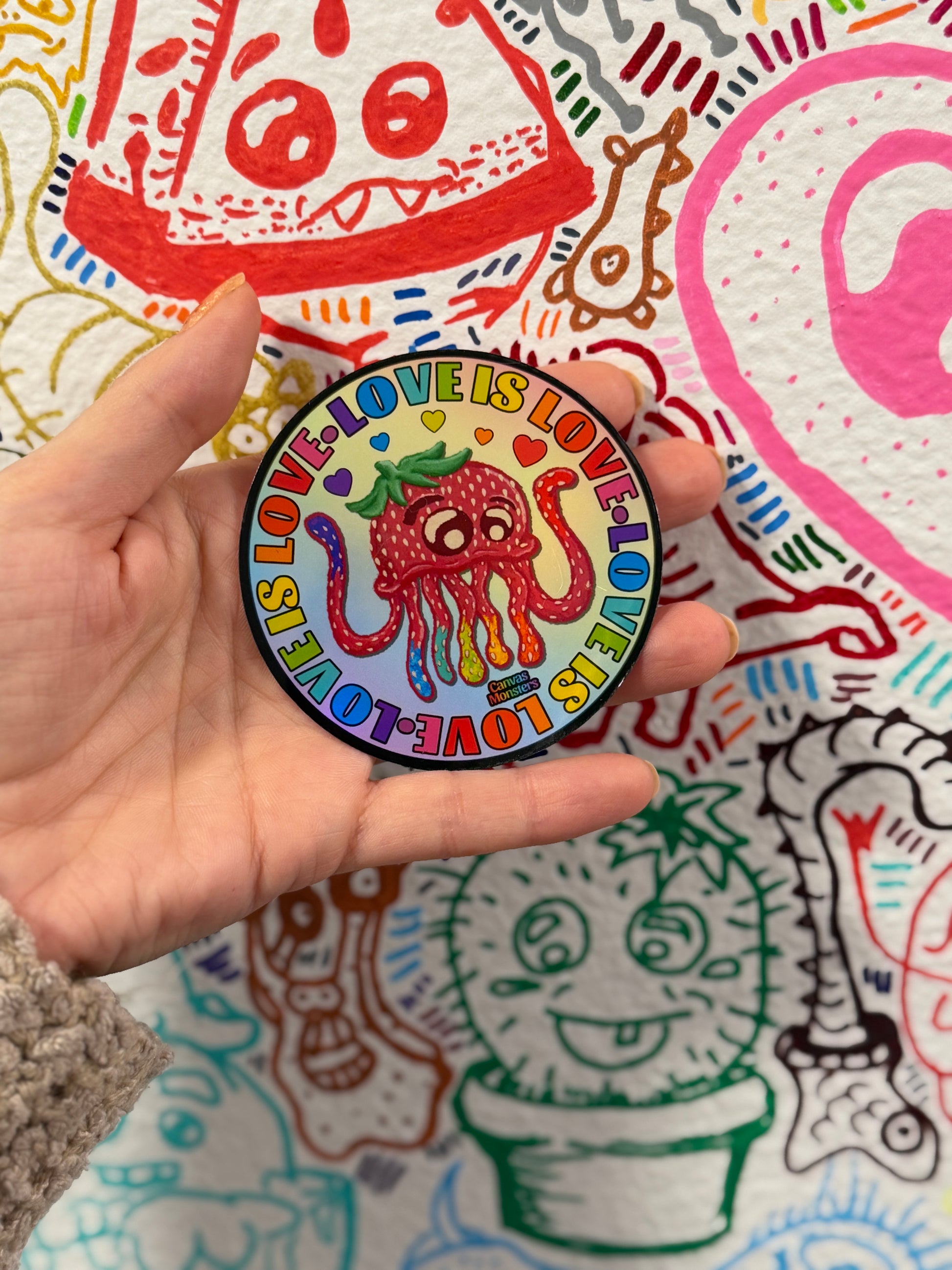 A hand holding a round 3-inch sticker featuring a colorful rainbow strawberry-octopus character with the text 'LOVE IS LOVE' repeated around it, set against a background of a hand-drawn, colorful illustrated pattern.
