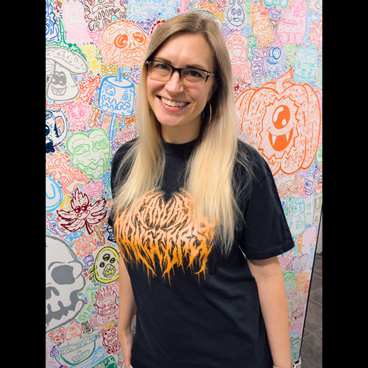 A smiling woman in front of a monster mural of all different colors and characters. She is wearing the black shirt with orange metal font that reads CANVAS MONSTERS that is featured in this product listing, 