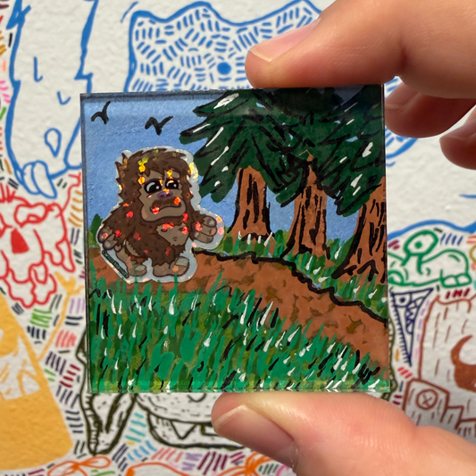 A hand holds a small square painted magnet featuring a brown furry creature walking through green grass, trees, and a blue sky. The background is a white surface covered in colorful doodles and sketches.