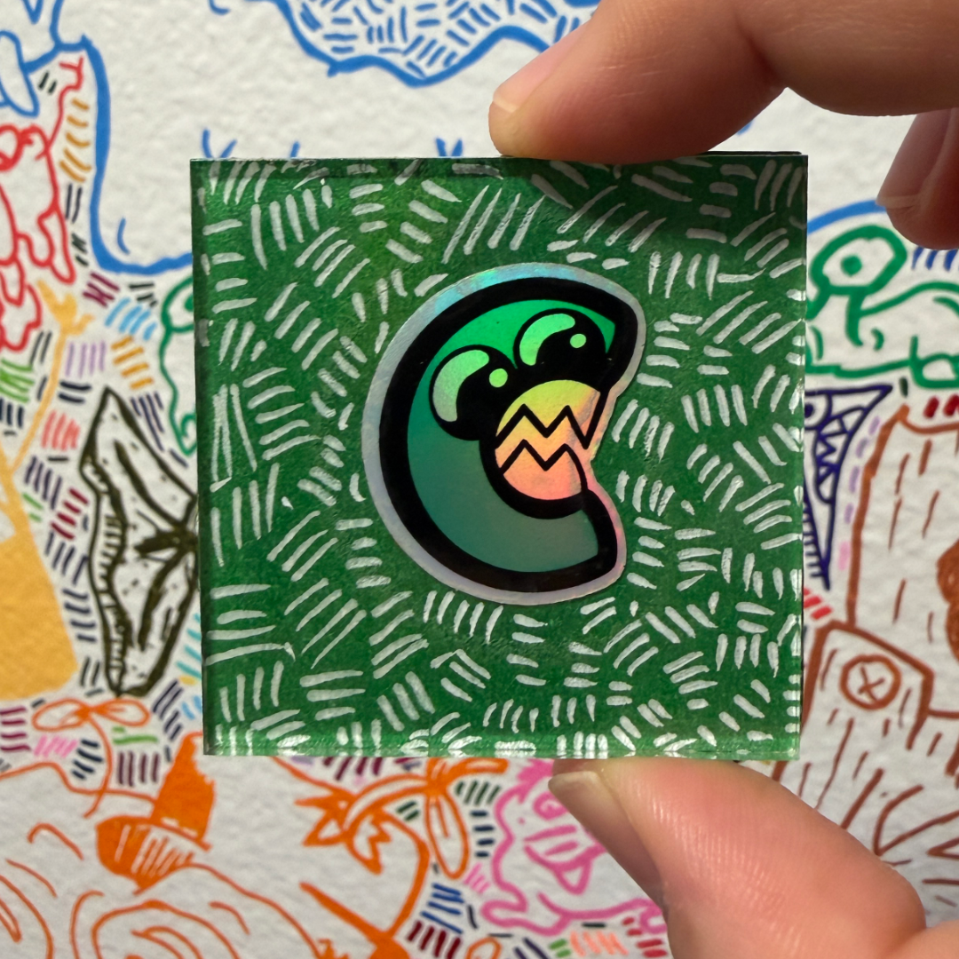 A hand holds a small square green magnet featuring Canvas Monsters C logo with a holographic sheen and is shown against a white background with colorful doodle-style illustrations.