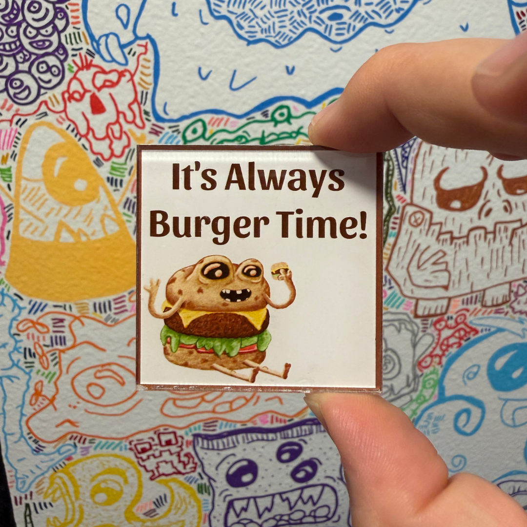 A hand holds a small square magnet that reads 'It's Always Burger Time!' with a cartoon burger character, against a colorful illustrated background.