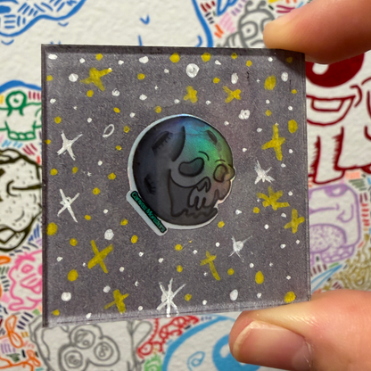A hand holds a square gray magnet featuring a skull shaped moon character with a holographic face, surrounded by white and yellow star-like speckles on a gray background.