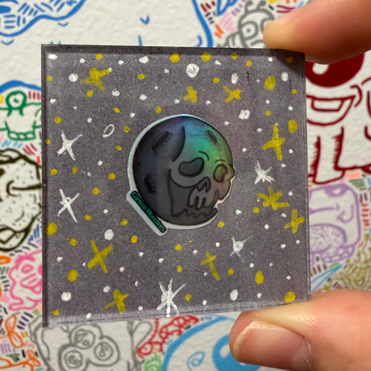 A hand holds a square gray magnet featuring a skull shaped moon character with a holographic face, surrounded by white and yellow star-like speckles on a gray background.