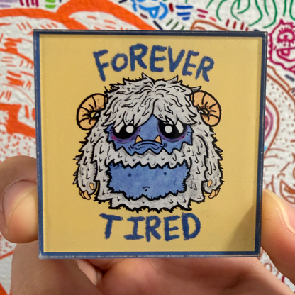 A hand holds a square magnet featuring a cartoon monster with the text 'FOREVER TIRED' in blue on a pale yellow background. The magnet has a dark blue border and is shown against a colorful patterned surface.