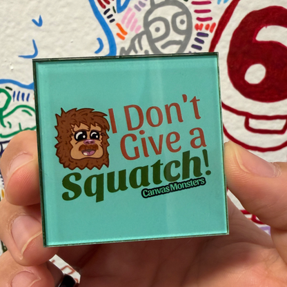 A hand holding a square acrylic magnet with a turquoise background. The sign features a cartoon Bigfoot-like creature and the text 'I Don't Give a Squatch!' in red and green lettering, with 'CanvasMonsters' written below.
