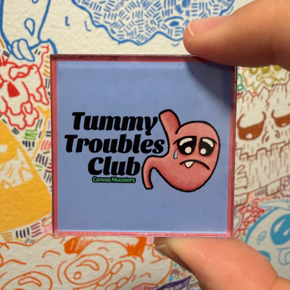 A square magnet featuring the text 'Tummy Troubles Club' and a cartoon crying stomach character, held by fingers against a colorful patterned background.