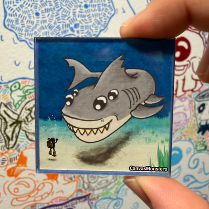 A hand holds a square magnet featuring a cartoon gray shark character swimming underwater, with a small figure on the left and the text 'CanvasMonsters' in the bottom right corner. The background shows a colorful, hand-drawn collage.