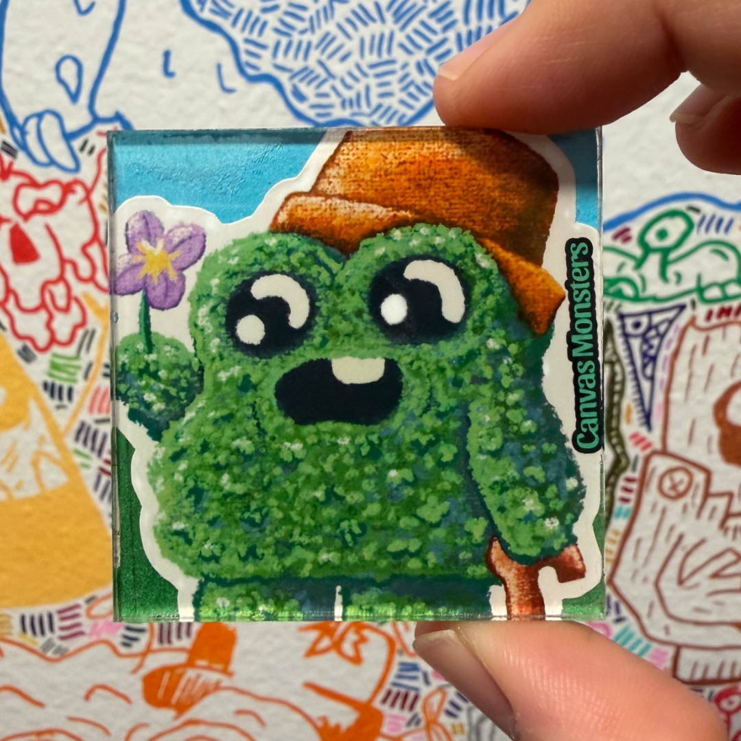 A hand holds a square magnet featuring a green cartoon monster wearing an orange hat and holding a purple flower.