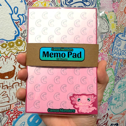 A hand holding a packaged memo pad from Canvas Monsters. The memo pad has a pink and white design with a repeating axolotl monster pattern, a brown paper band label, and a pink monster illustration in the bottom right corner.
