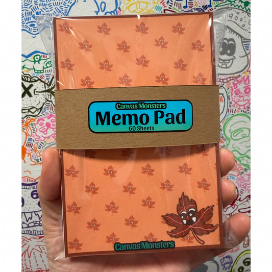 A hand holding a packaged orange memo pad with a brown paper band label that reads 'Canvas Monsters Memo Pad 60 Sheets.' The pad has a repeating leaf pattern and a cartoon leaf graphic near the bottom, photographed against a colorful illustrated background.