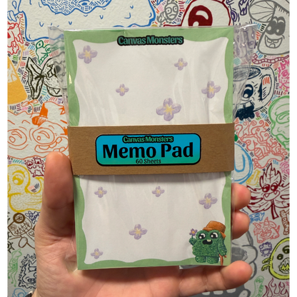A hand holding a package of 4"x6" Canvas Monsters Memo Pad 60 sheets, featuring a white paper pad with light purple flower patterns and a brown paper band label, set against a colorful, doodle-filled background.