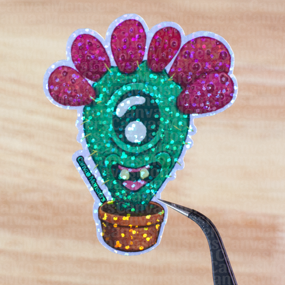 Glitter sticker of a cactus character with red pears, held above a wooden surface