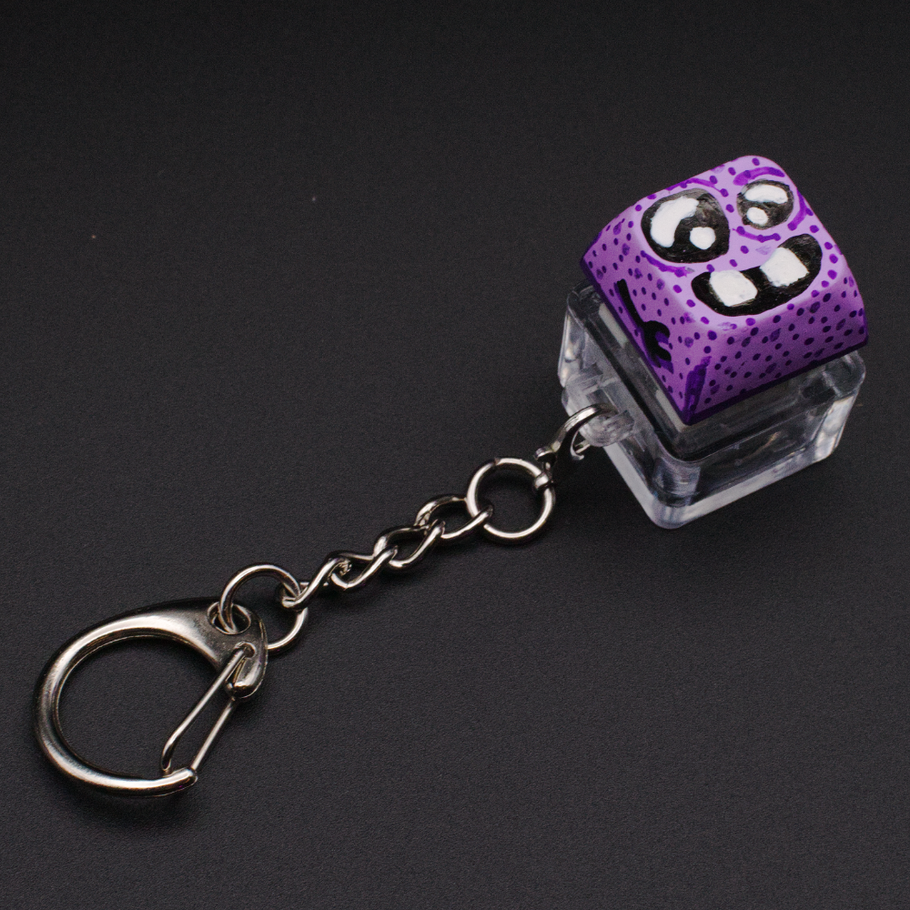 Photo of a key cap keychain on a black background. Cap is painted with a purple monster character.