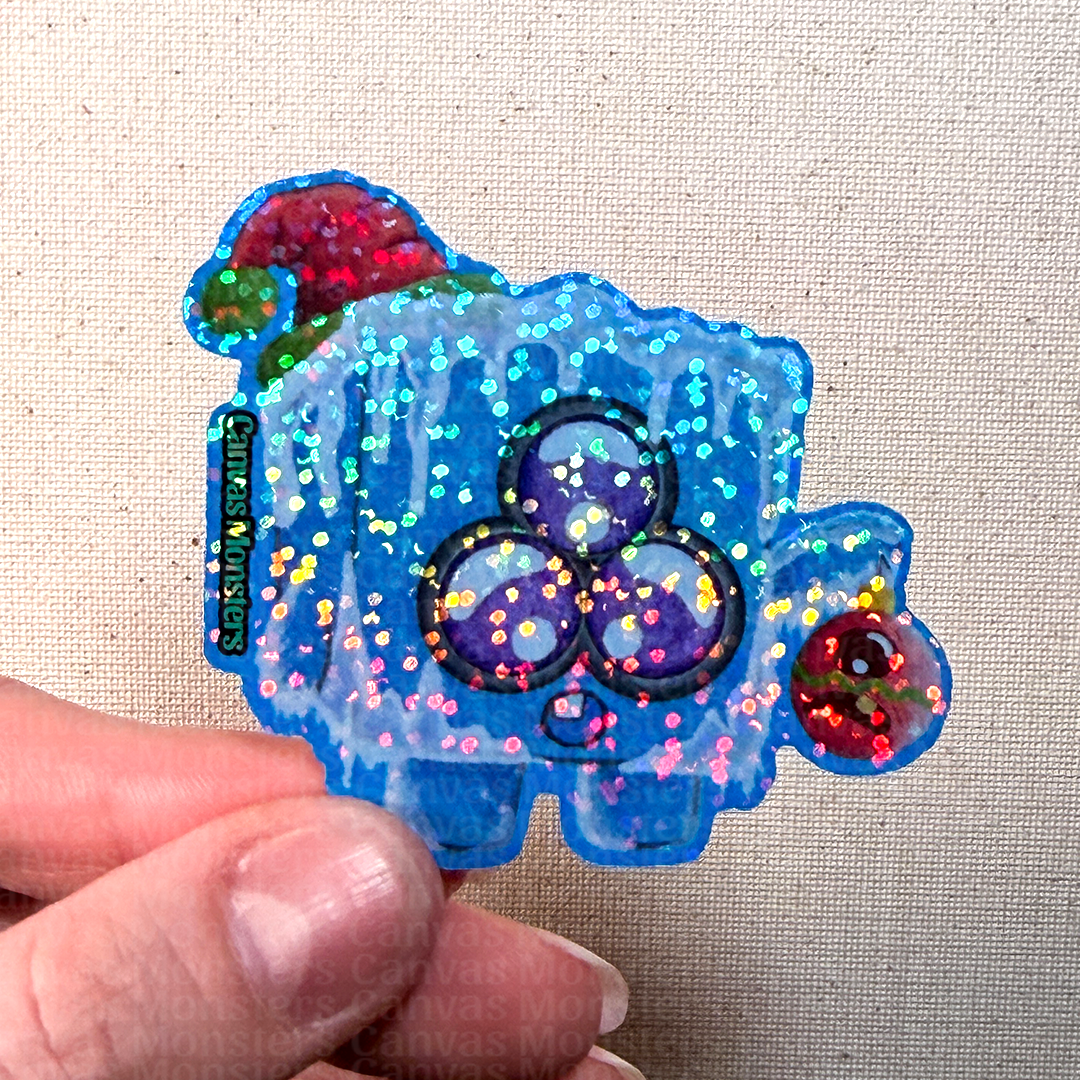 Colorful glittery sticker of an ice cube character held by a hand on a beige fabric background