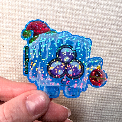 Colorful glittery sticker of an ice cube character held by a hand on a beige fabric background