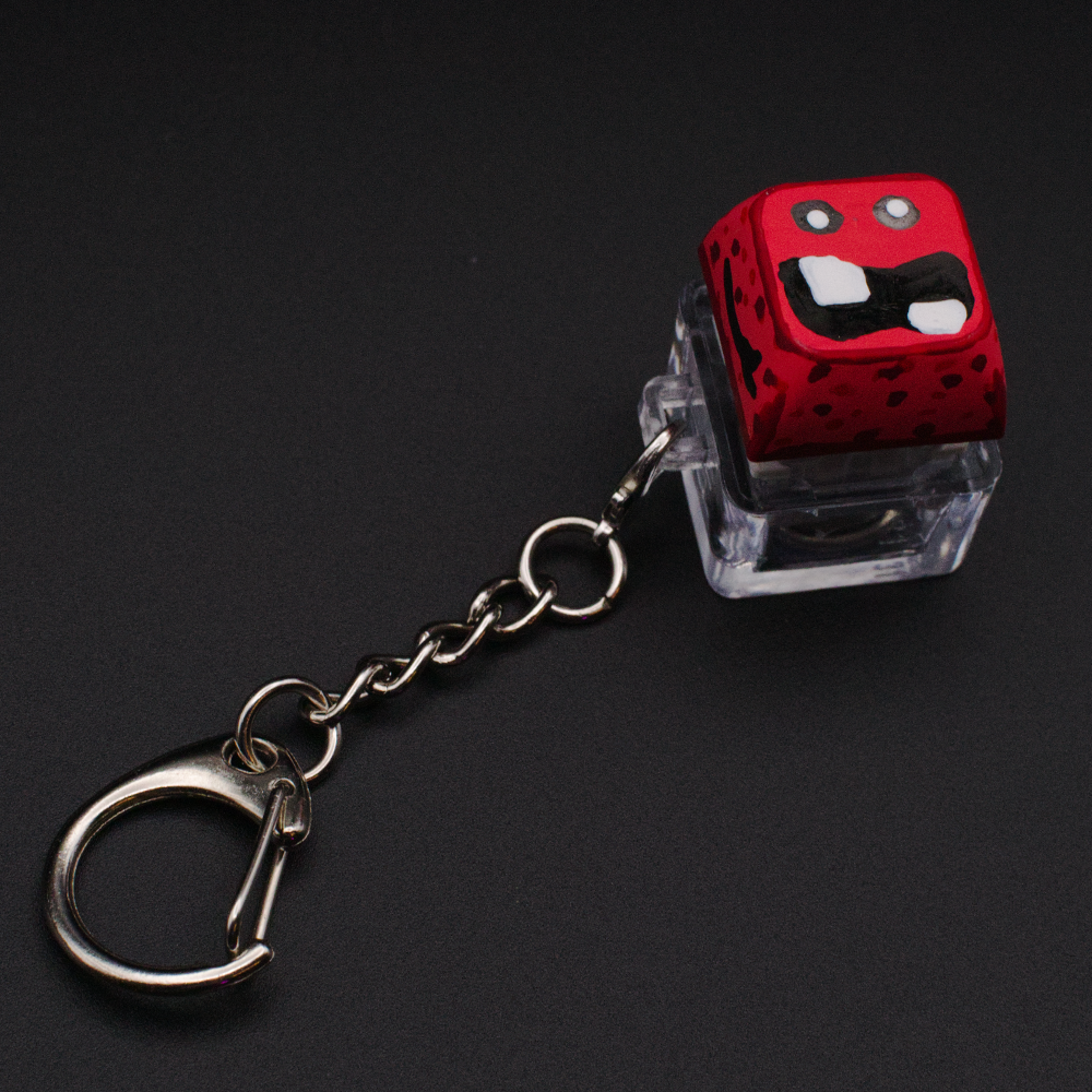 Photo of a key cap keychain on a black background. Cap is painted with a red monster character.