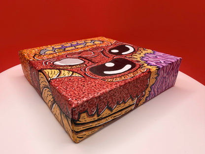 Red and gold monster character painted on a canvas block with purple brains on a red and white background.