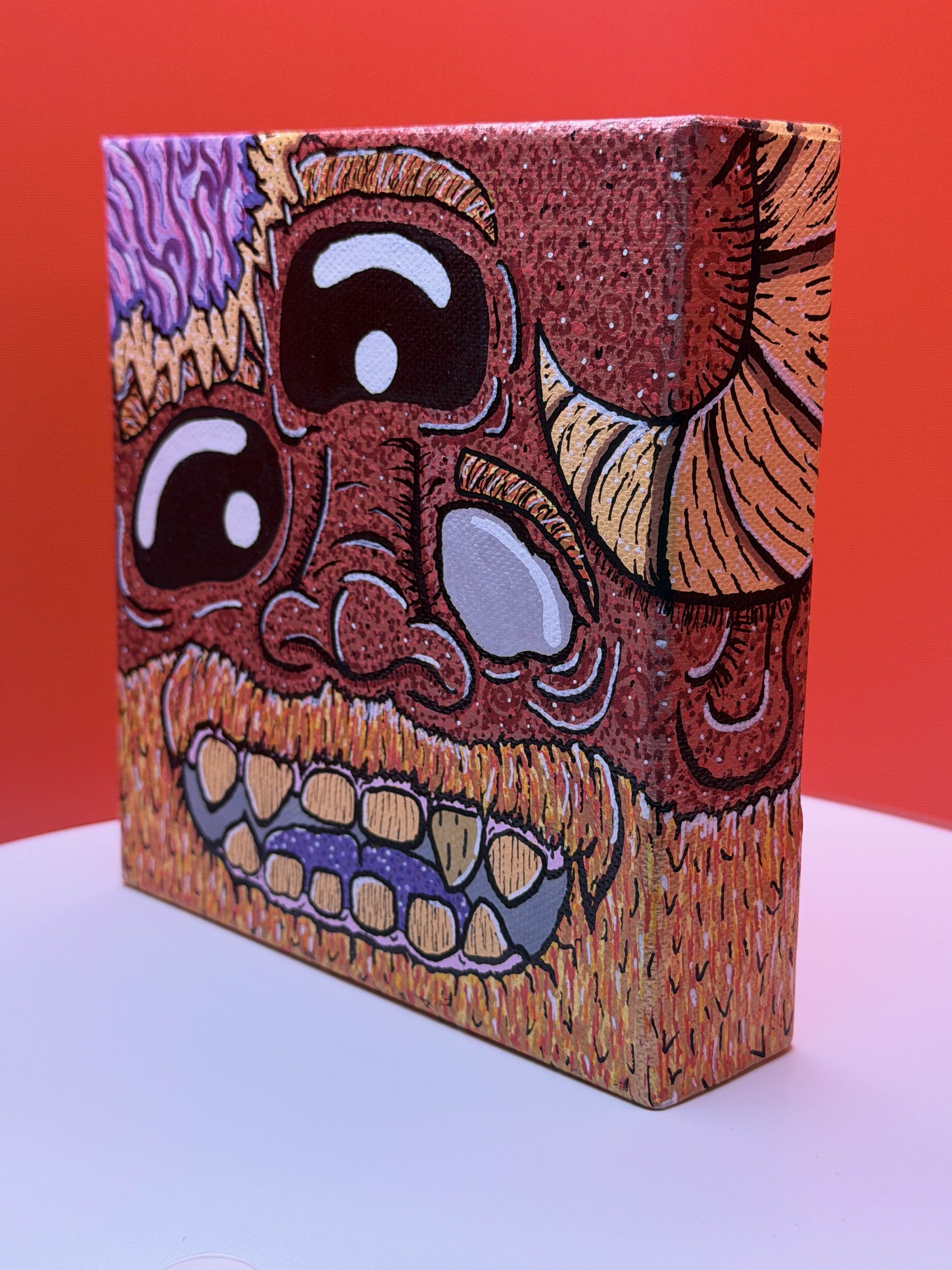 Red and gold monster character painted on a canvas block with purple brains on a red and white background.
