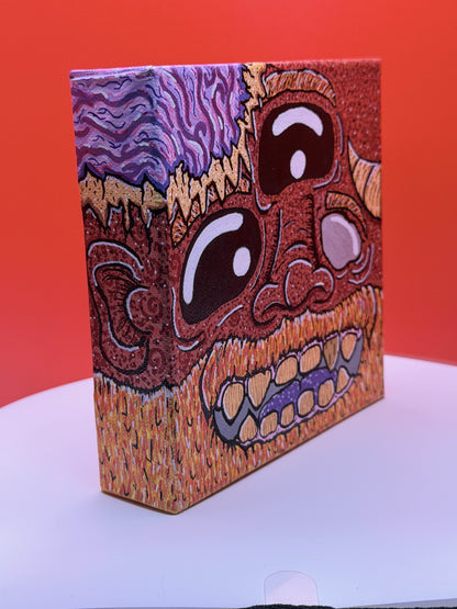 Side view of a red and gold monster character painted on a canvas block with purple brains on a red and white background.