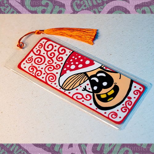 White paper bookmark with Canvas Monsters character "Shroomi" painted on it with a red swirl background and border, in a plastic sleeve and matching red tassel.