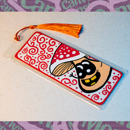 White paper bookmark with Canvas Monsters character "Shroomi" painted on it with a red swirl background and border, in a plastic sleeve and matching red tassel.