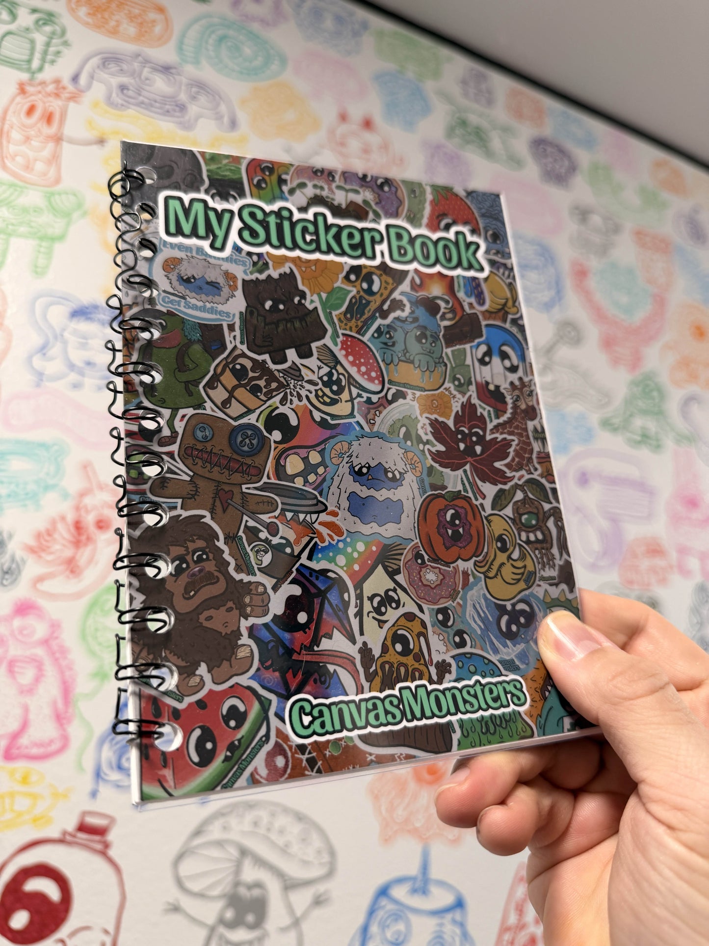 Hand holding a bound book with the title "My Sticker Book" and "Canvas Monsters" on the footer.