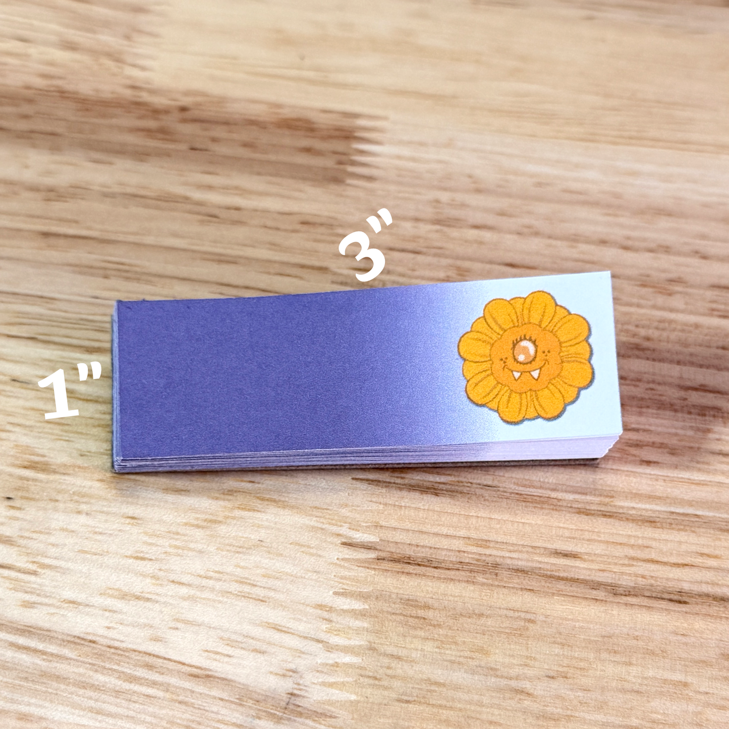 1x3 inch sticky note pad with a purple gradiant and a cartoon sunflower on the right side, sitting on a wooden background.