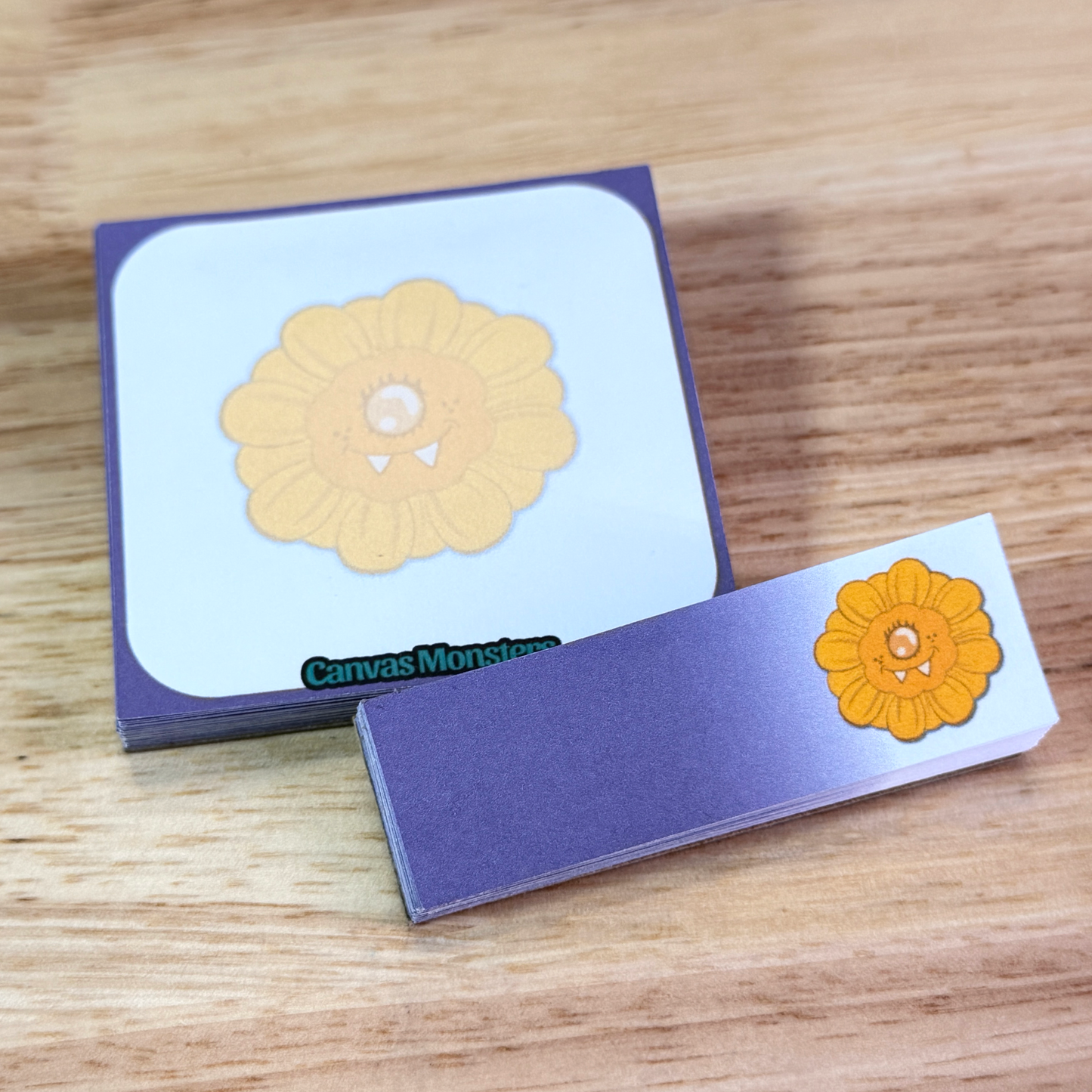 A set of matching sticky notepads, with purple and white, with a cartoon sunflowers character, sitting on a wooden background.