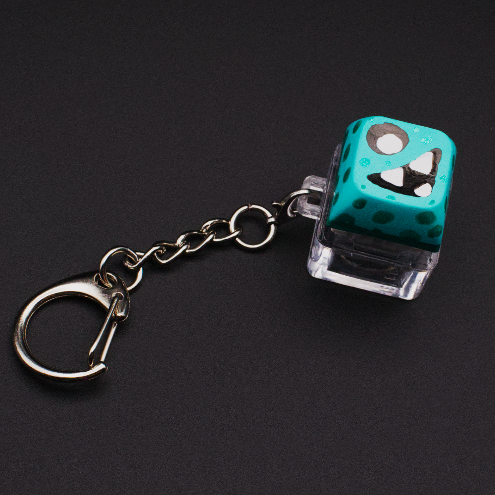 Photo of a key cap keychain on a black background. Cap is painted with a light blue monster character.