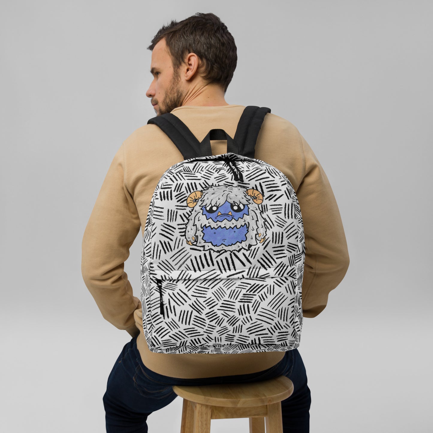 A man wearing a beige sweatshirt and dark jeans sits on a wooden stool, viewed from behind, wearing a white backpack with a black geometric pattern and a blue monster graphic on the back.