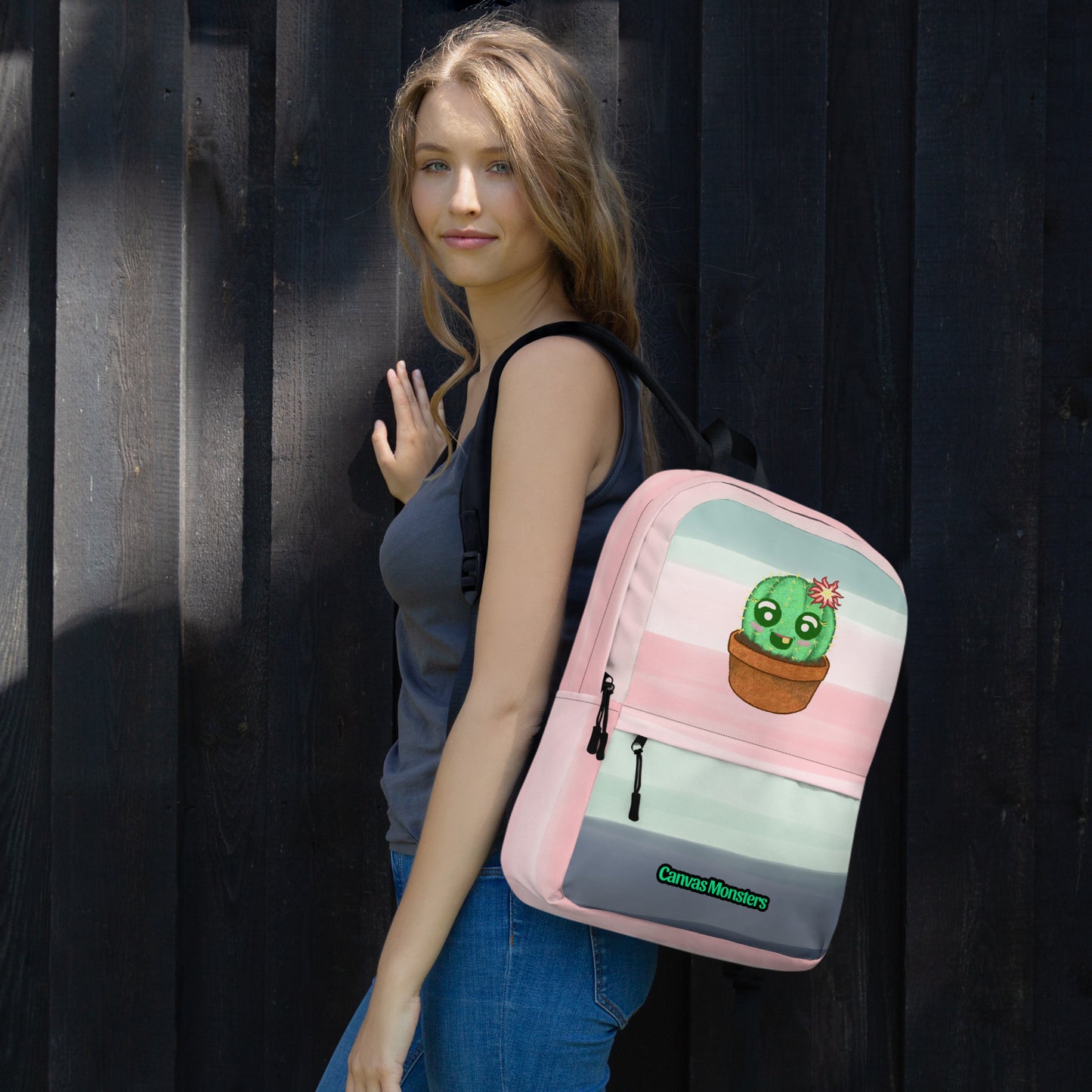 A woman wearing a gray tank top and blue jeans carries a pink backpack with a cute cartoon cactus in a pot on the front, standing against a dark wooden plank wall.