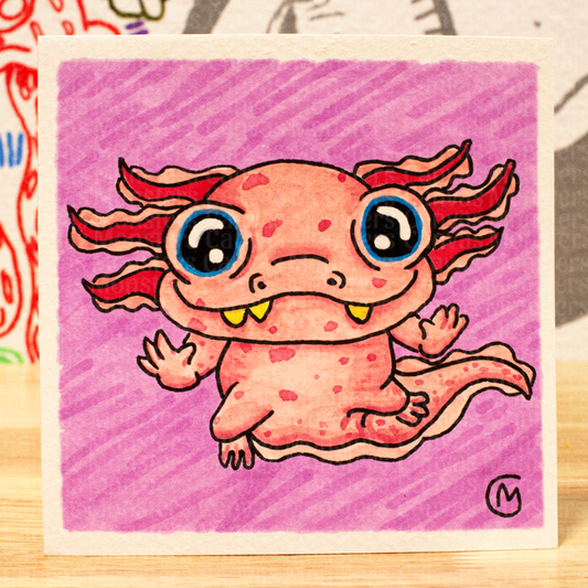 A square art print or card featuring a cute pink cartoon axolotl on a purple background, photographed on a wooden surface with colorful objects blurred in the background.