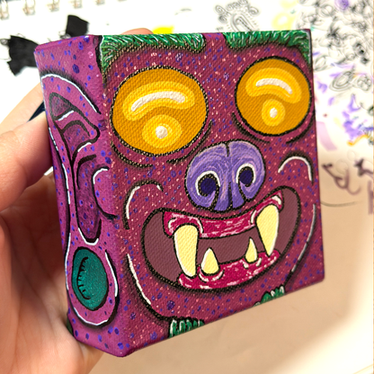 an up close picture of a monster block painting. This monster is featured with yellow eyes, a purple nose, burgundy skin, yellow teeth, and on the side of the canvas block, we see the ear and the earring of this monster. 