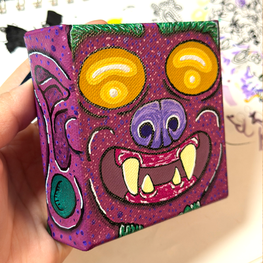 an up close picture of a monster block painting. This monster is featured with yellow eyes, a purple nose, burgundy skin, yellow teeth, and on the side of the canvas block, we see the ear and the earring of this monster. 