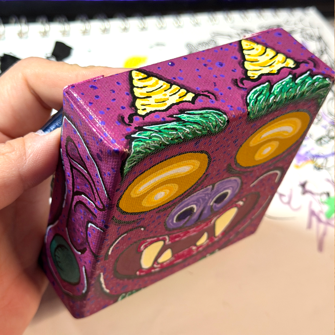 an up close picture of a monster block painting. This monster is featured with yellow eyes, a purple nose, burgundy skin, yellow teeth, and on the side of the canvas block, we see the ear and the earring of this monster.  This is a top down angle of the monster, showcasing it's horns. 