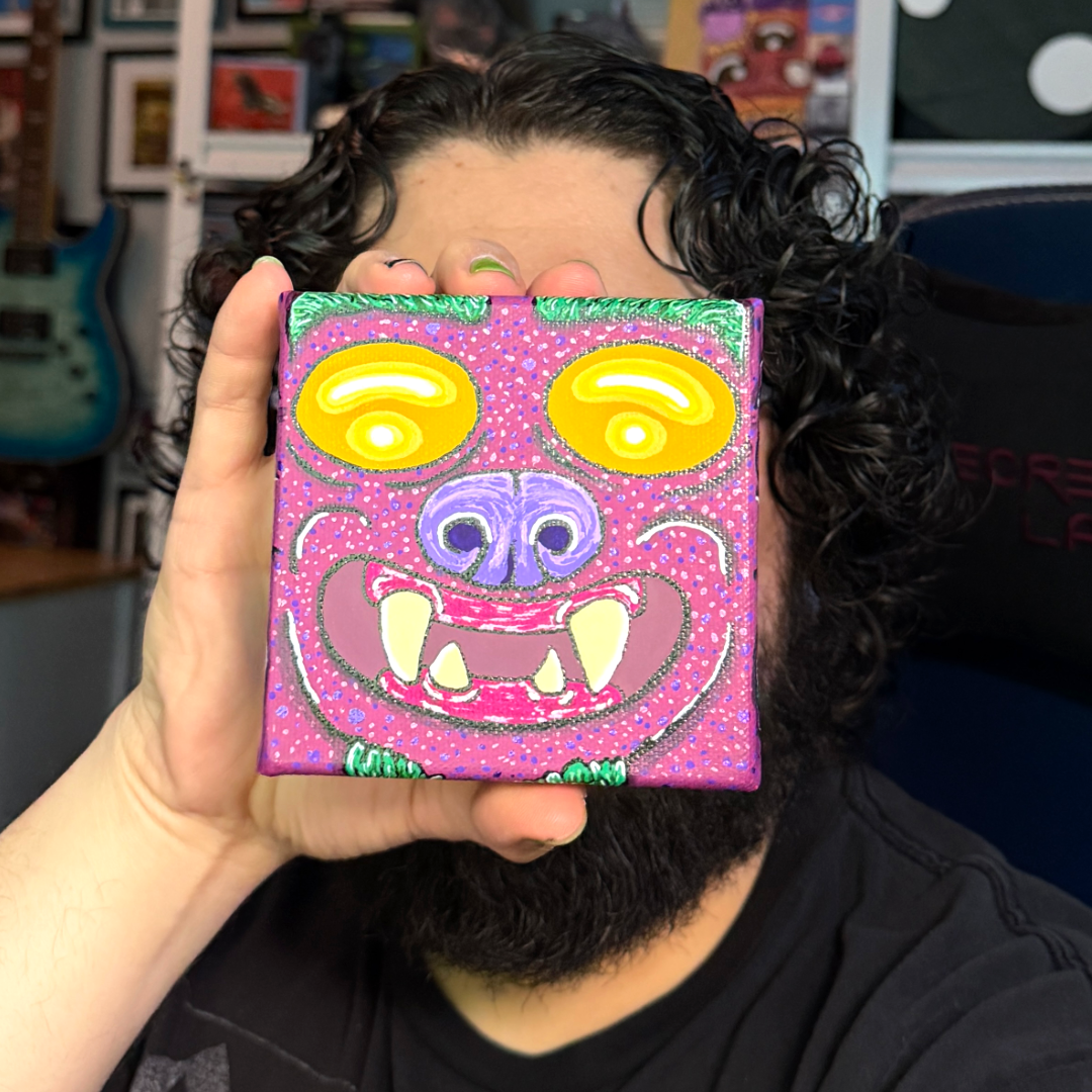 an up close picture of a monster block painting. This monster is featured with yellow eyes, a purple nose, burgundy skin, yellow teeth, and on the side of the canvas block, we see the ear and the earring of this monster.  This is a face on shot of the painting behind held by the artist Canvas Monsters. 