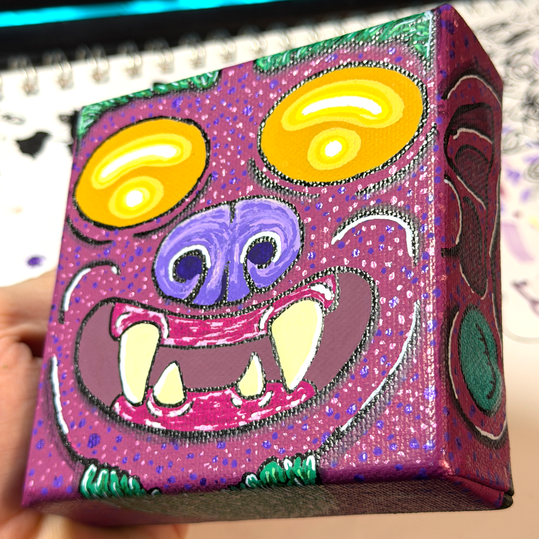 an up close picture of a monster block painting. This monster is featured with yellow eyes, a purple nose, burgundy skin, yellow teeth, and on the side of the canvas block, we see the ear and the earring of this monster.  This is a bottom up angle, we can see some of it's chin hair. 