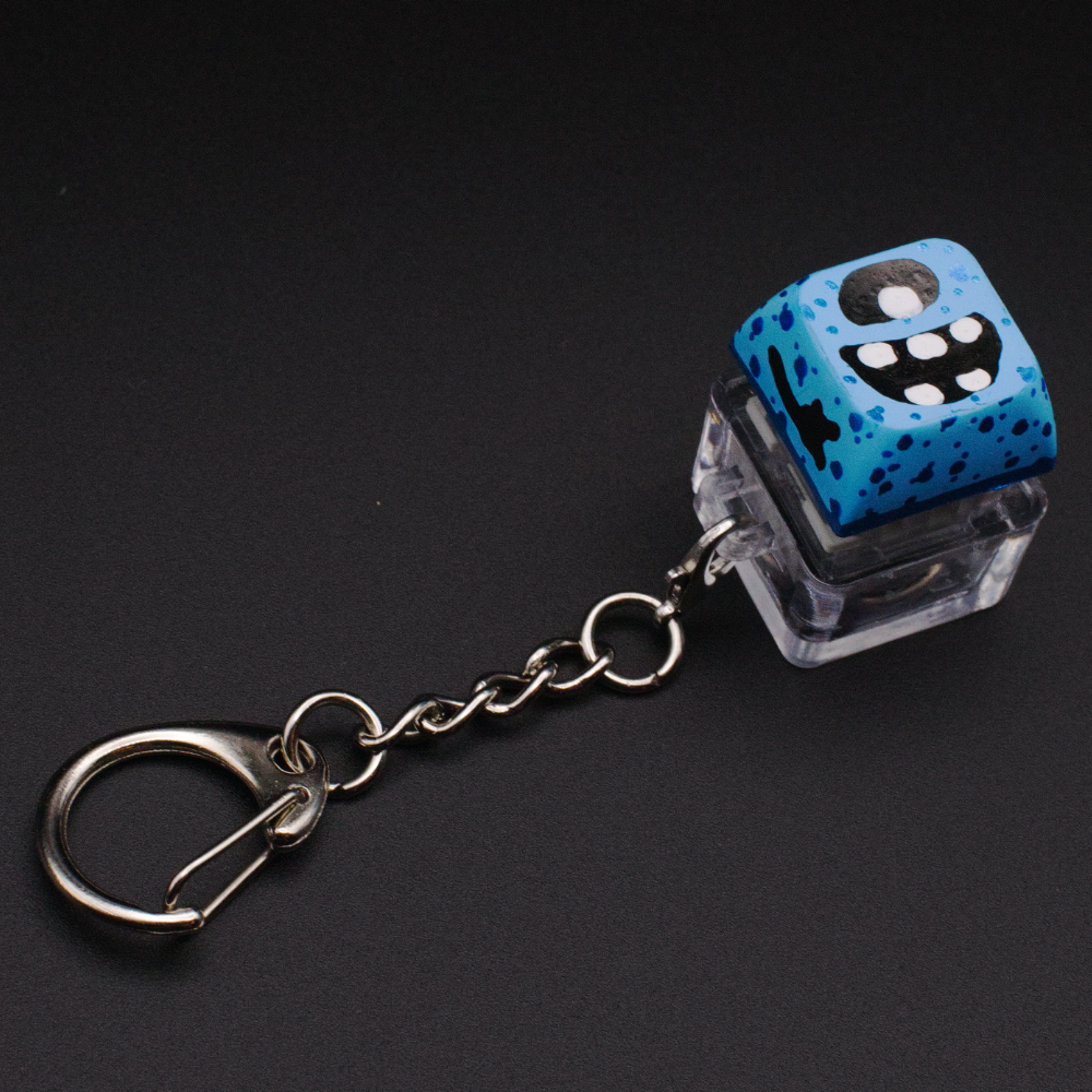 Photo of a key cap keychain on a black background. Cap is painted with a blue monster character.