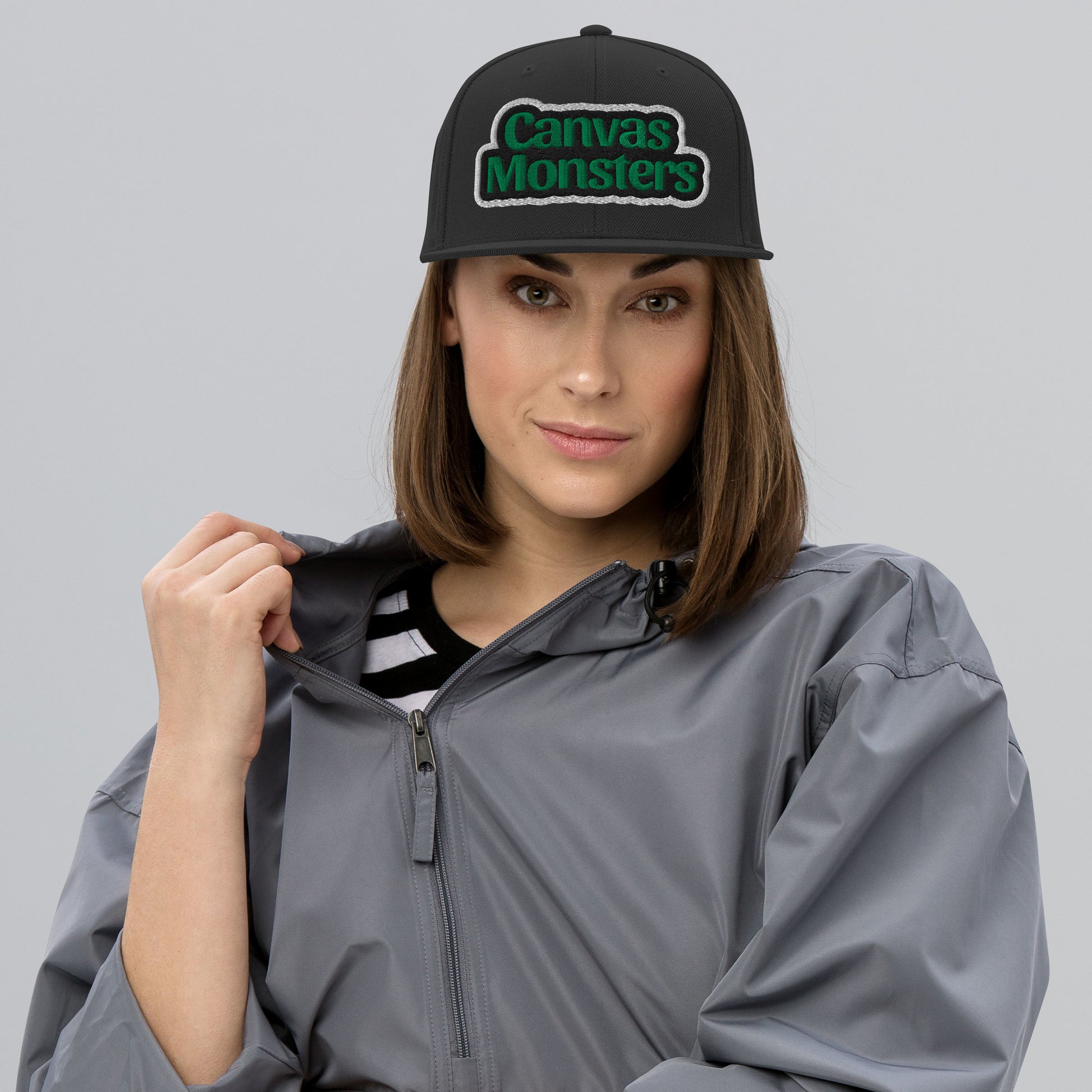 Woman wearing a gray raincoat and a black cap with 'Canvas Monsters' logo on a gray background