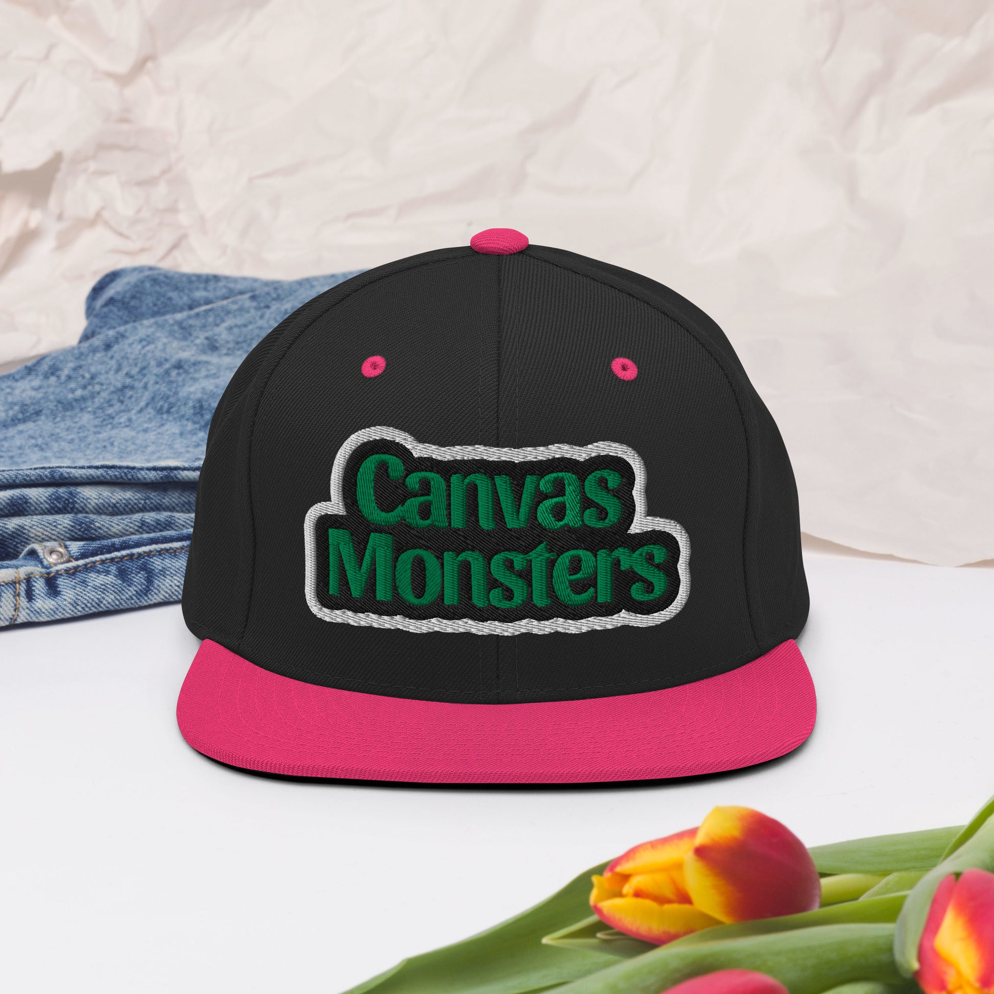 Black and pink cap with 'Canvas Monsters' logo on a light background with denim and tulips.