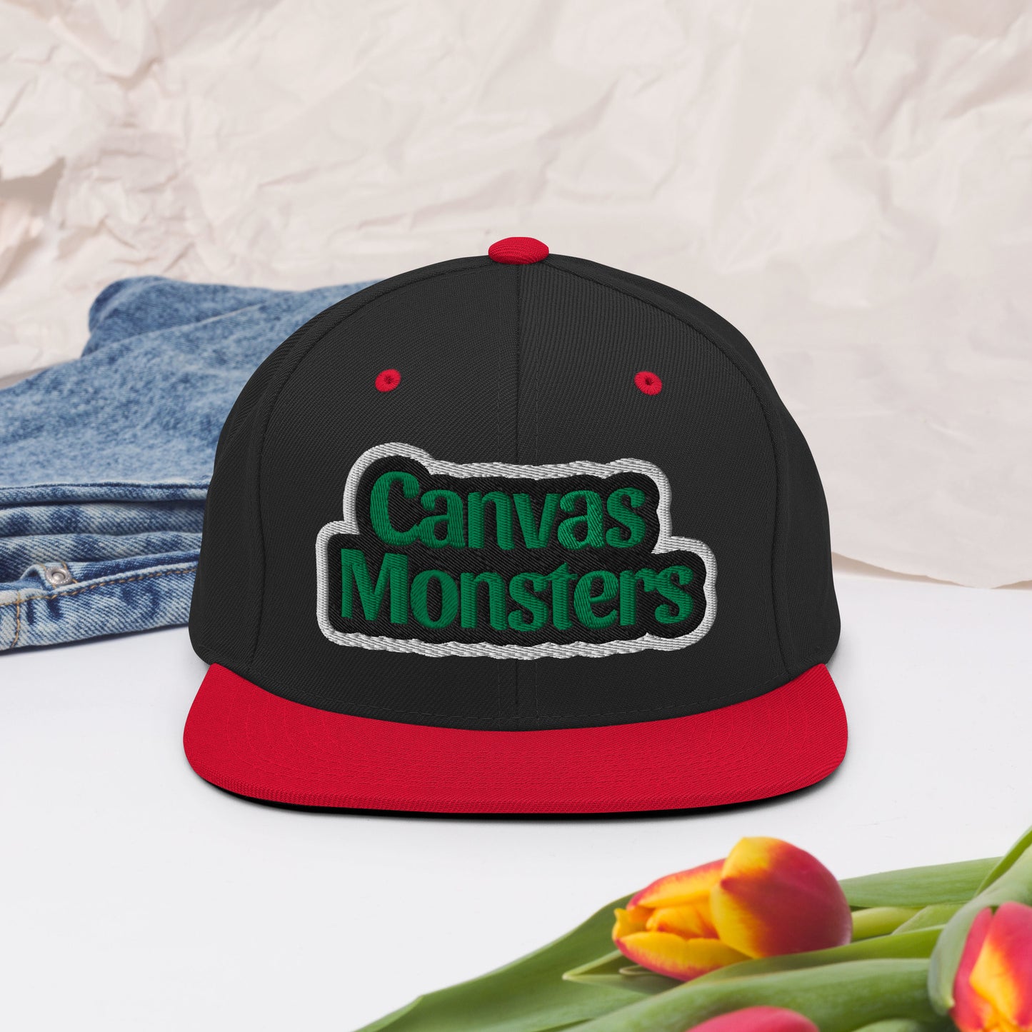 Black and red cap with 'Canvas Monsters' logo on a light background with denim and flowers.