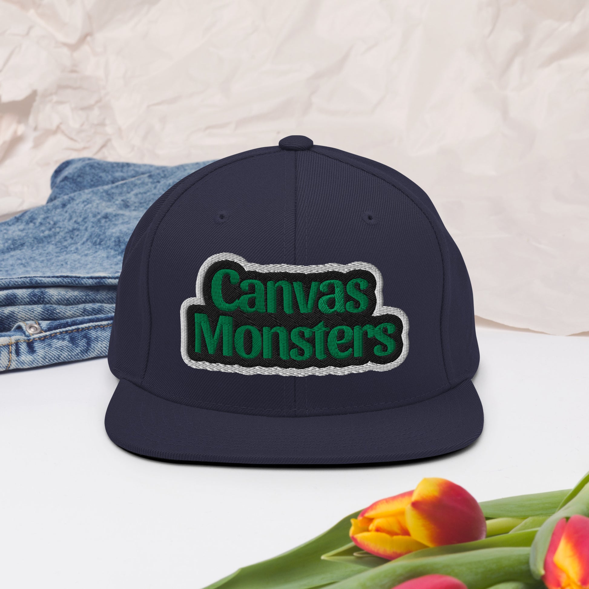 Navy blue cap with 'Canvas Monsters' logo on a light background with denim and tulips.