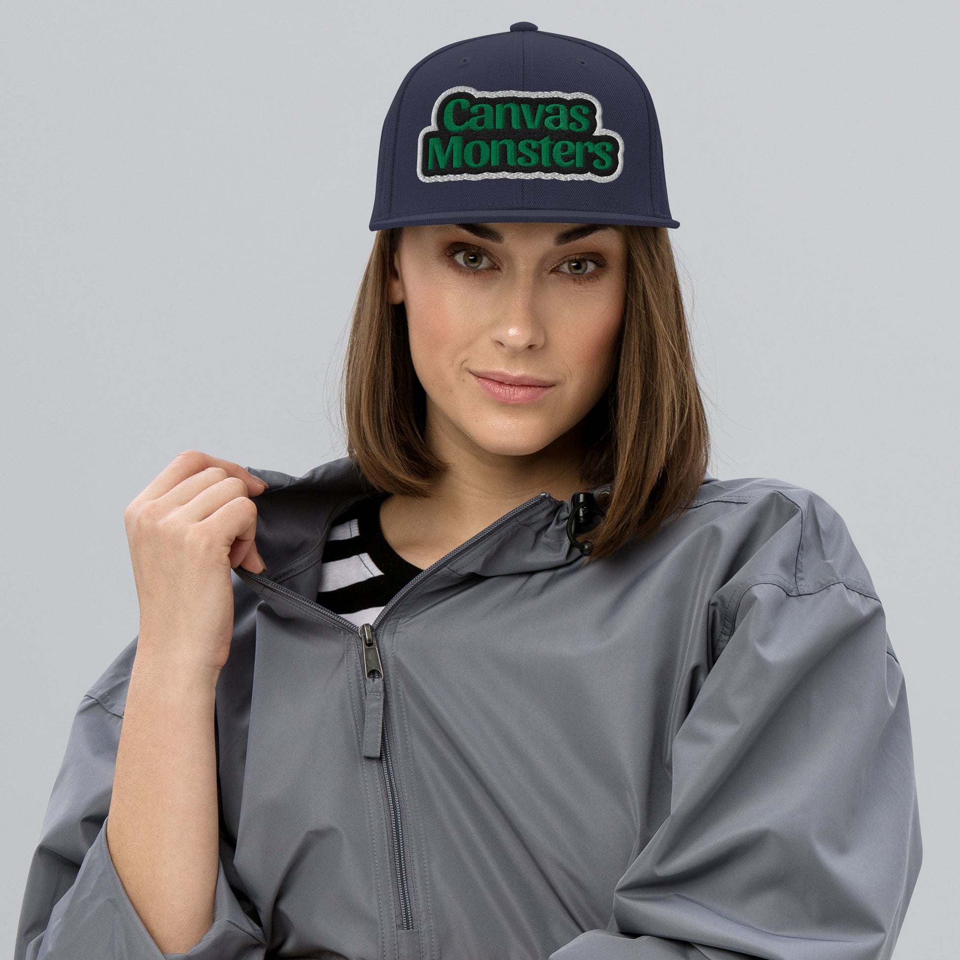 Woman wearing a gray raincoat and 'Canvas Monsters' cap on a gray background