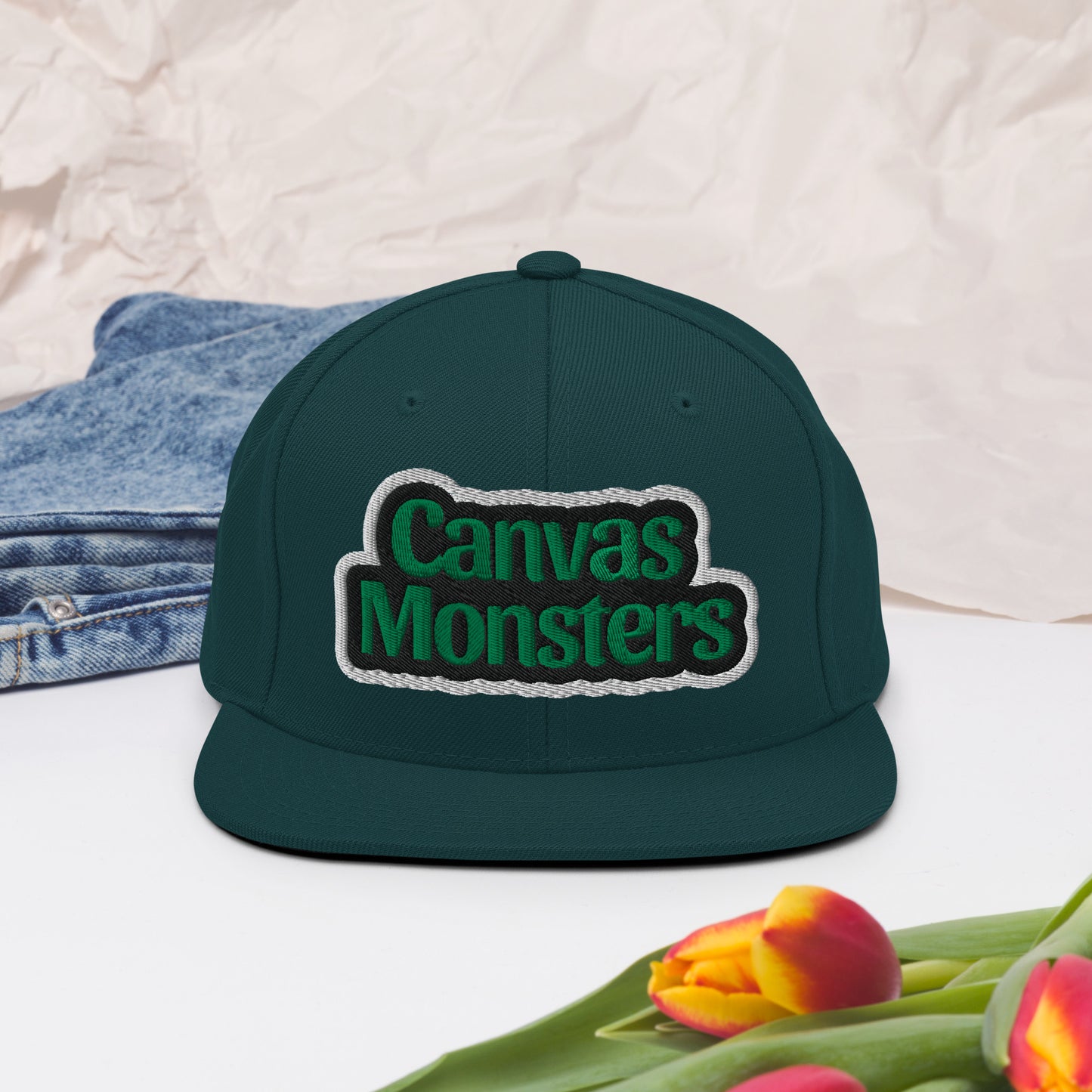 Green cap with 'Canvas Monsters' logo on a white surface with denim and tulips.