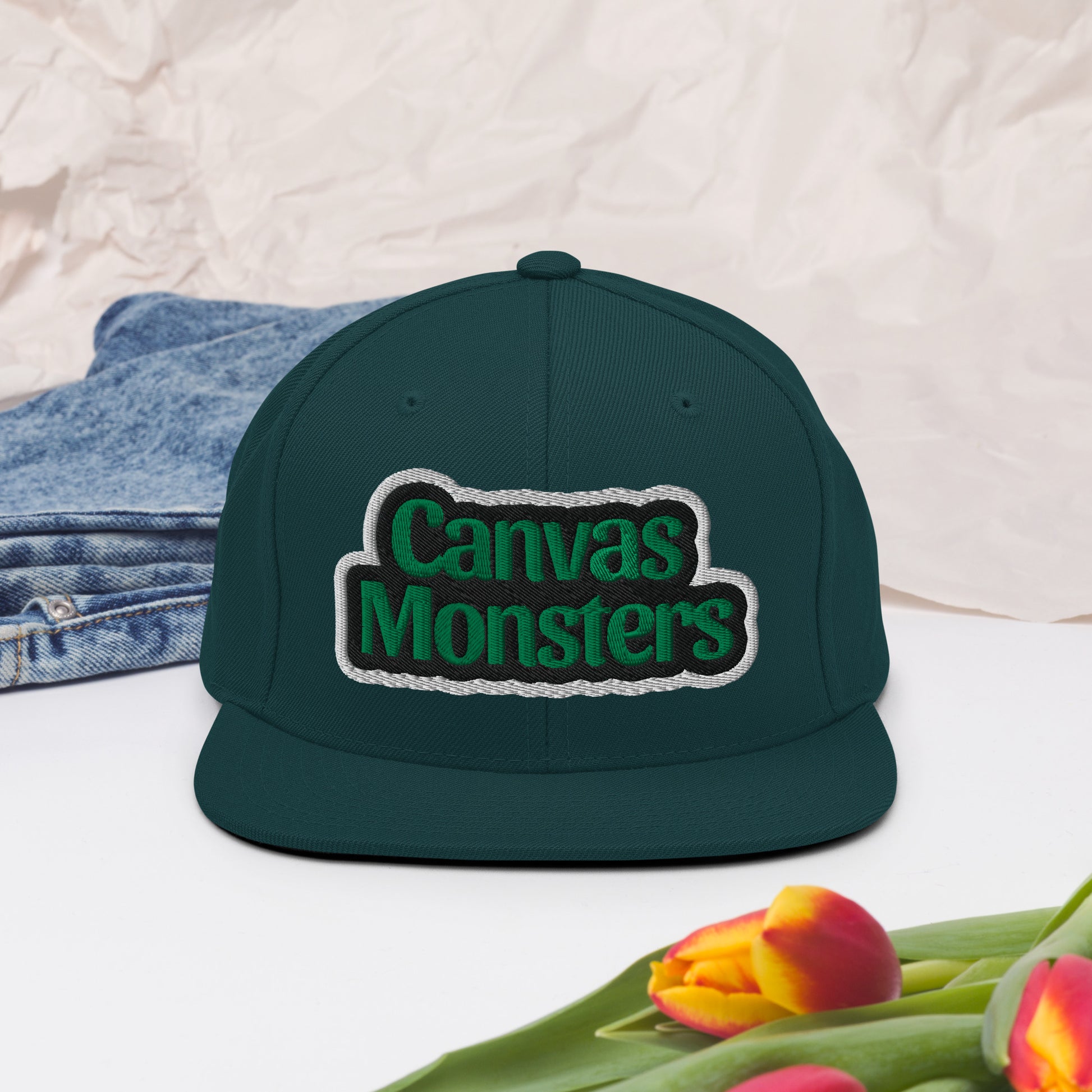 Green cap with 'Canvas Monsters' logo on a white surface with denim and tulips.