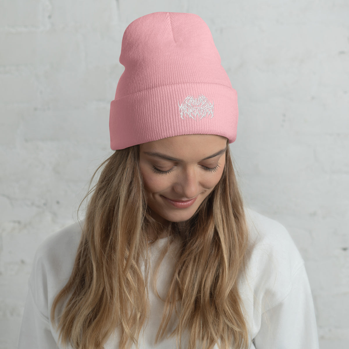 Woman with long blonde hair wearing a light pink knit beanie with a metal text logo on the front.