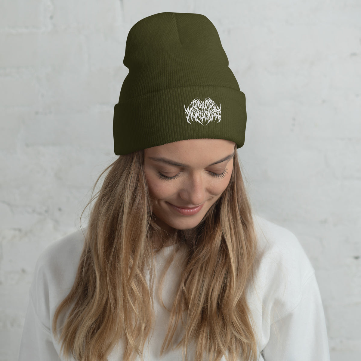 Woman with long blonde hair wearing an olive green knit beanie with a metal text logo on the front.