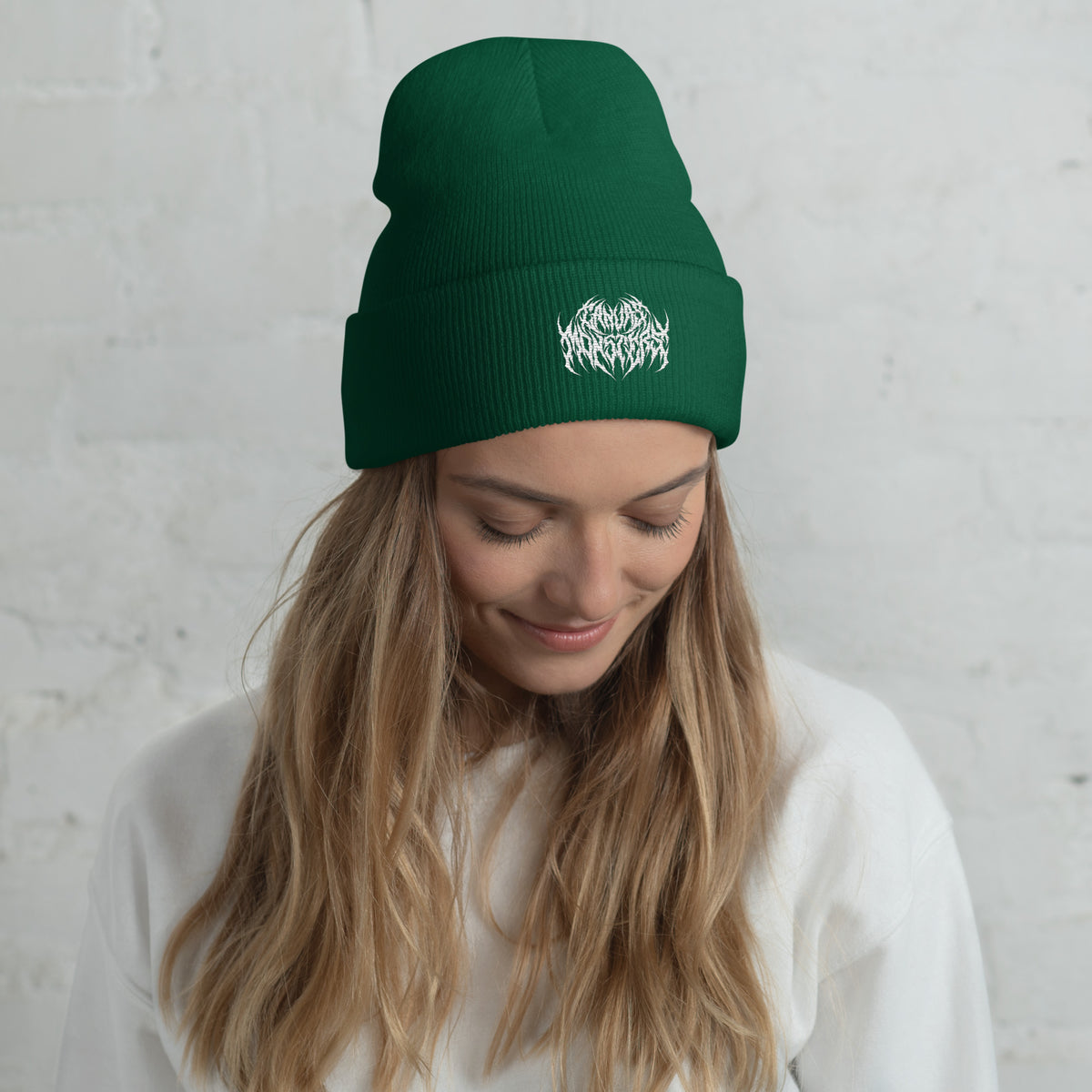 Woman with long blonde hair wearing a green knit beanie with a metal text logo on the front.
