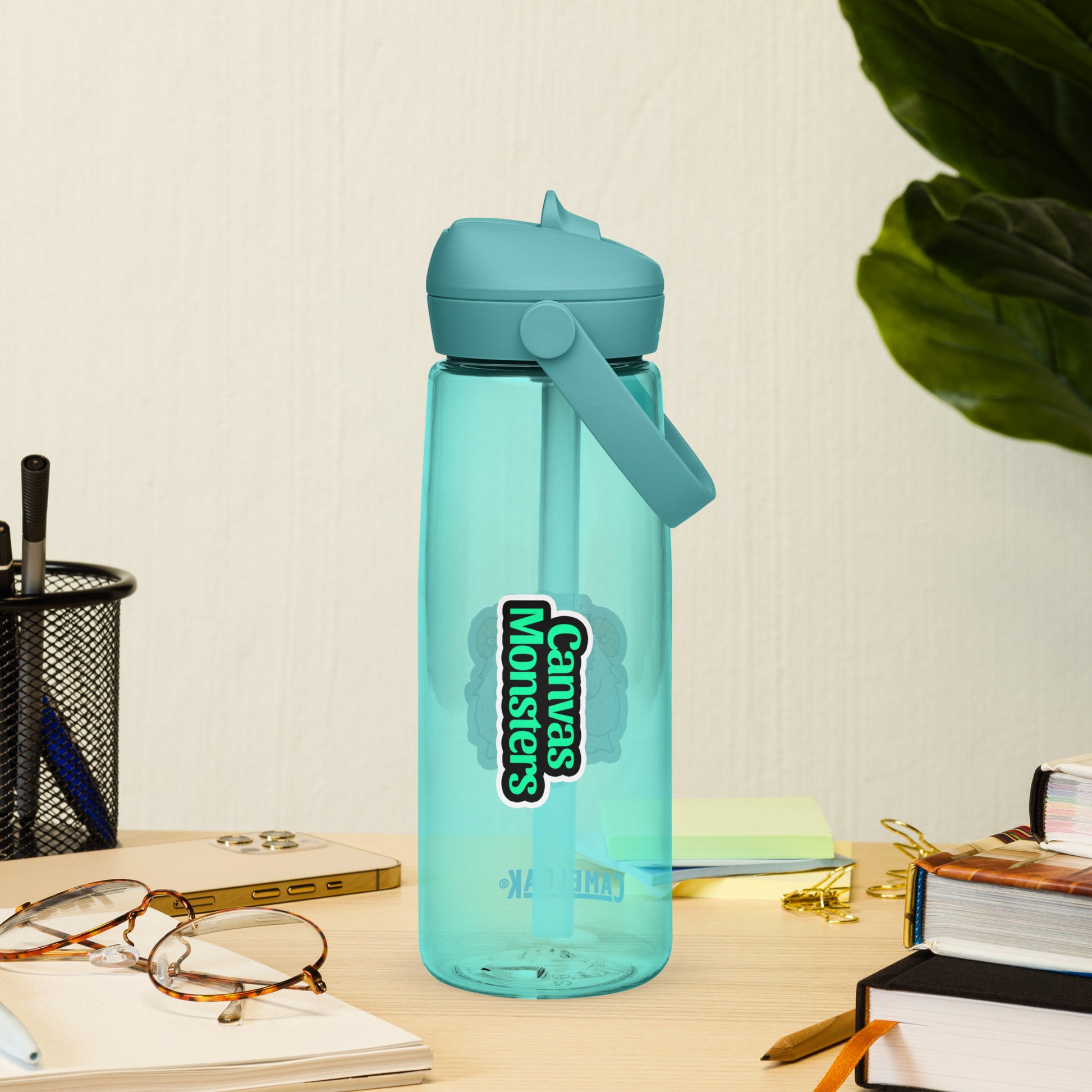 Blue water bottle with 'Canvases Monsters' logo on a desk with glasses and books.